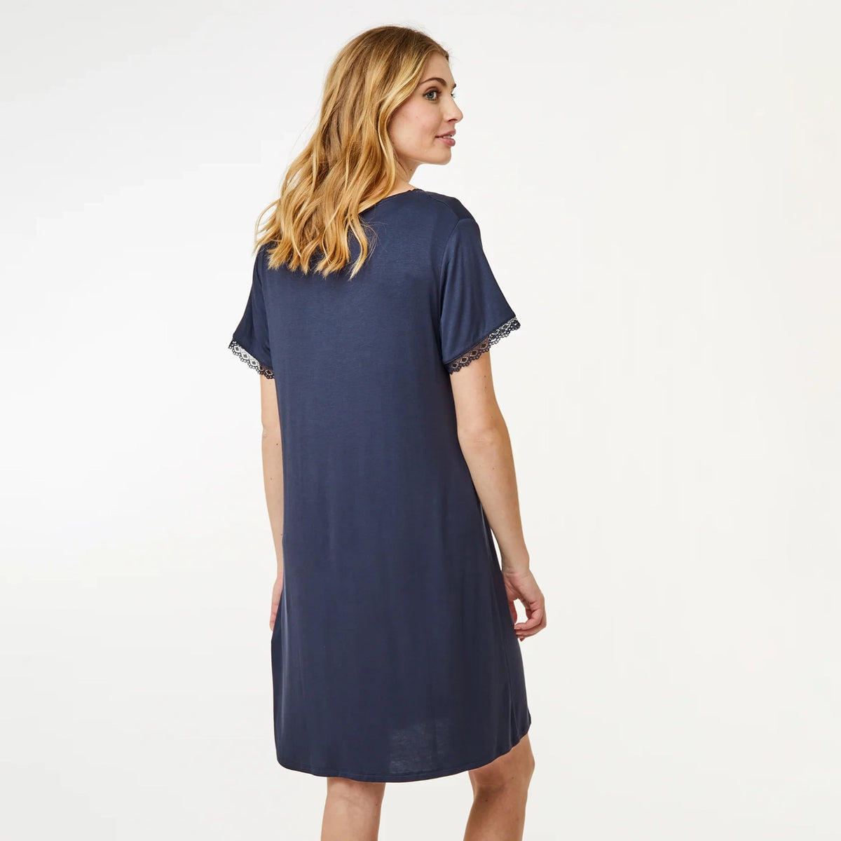 A woman wearing a navy blue short-sleeve nightshirt dress with lace trim on the sleeves, shown from a back-three-quarter view against a plain white studio background.