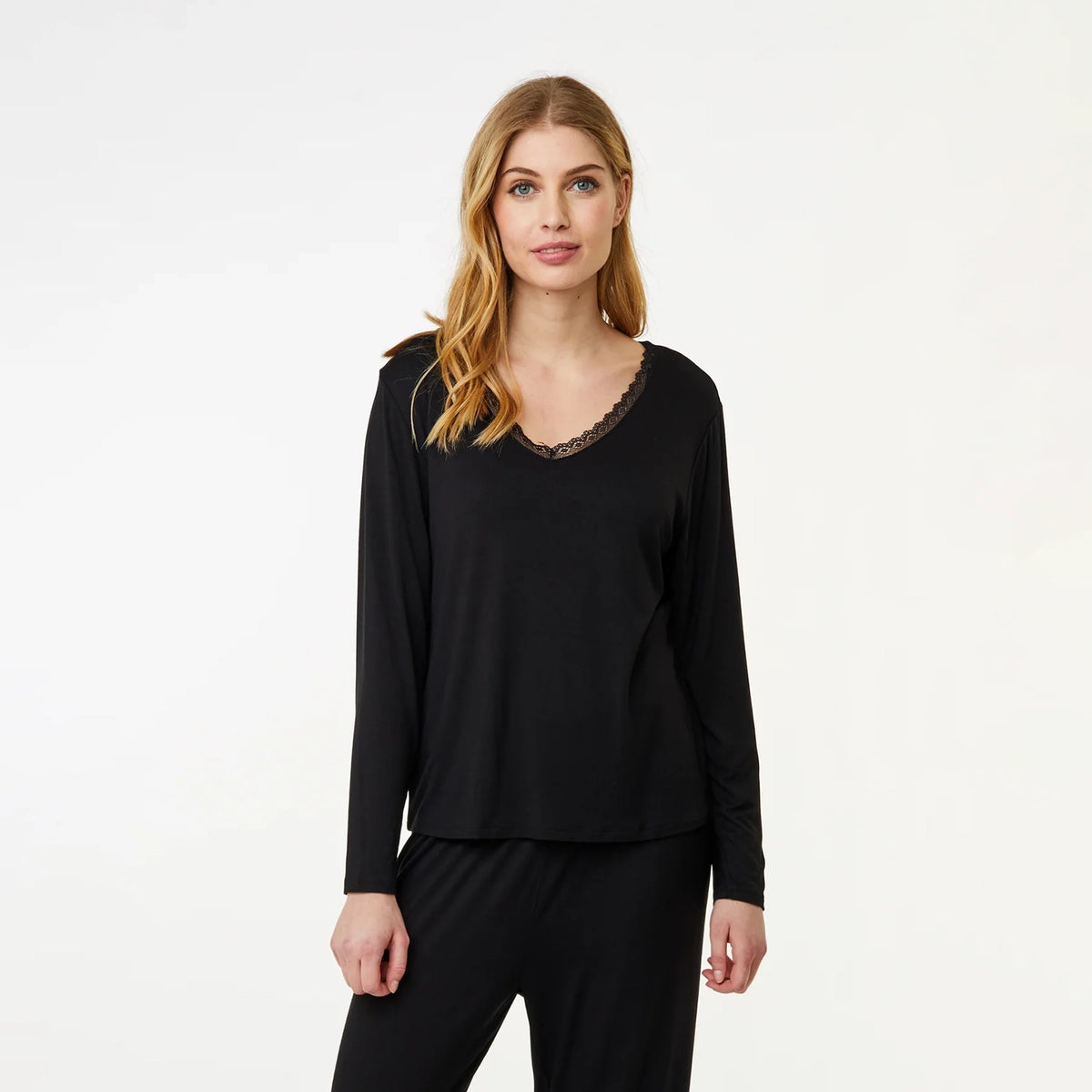 A woman modeling a black long-sleeve top with a scalloped lace V-neckline, paired with black pants, against a plain white studio background.