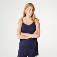 A woman modeling a navy blue camisole top with delicate lace trim at the neckline and hem, paired with matching navy pajama shorts, against a plain light gray studio background.