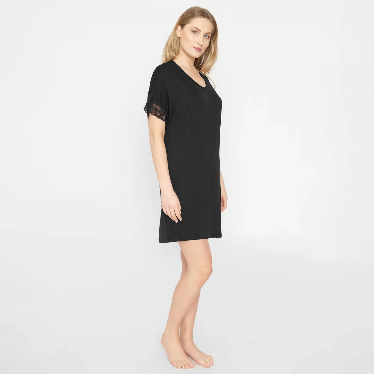A woman modeling a black short-sleeve nightgown with black lace trim on the sleeves, standing against a plain white studio background.