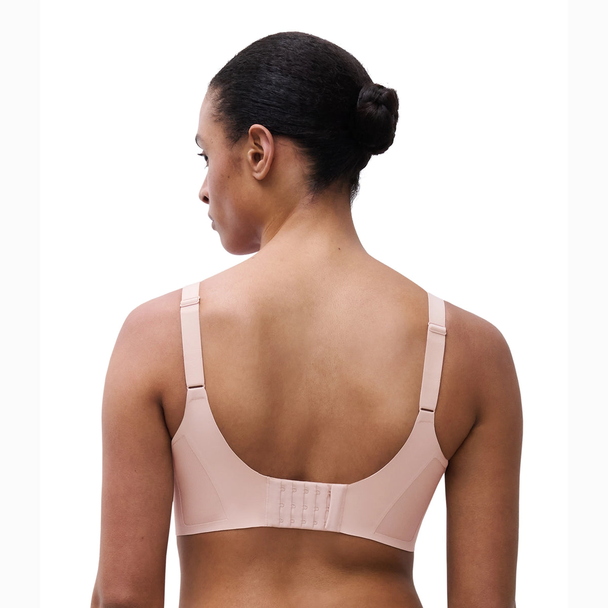 Rear view of a woman wearing a light pink seamless bra with adjustable shoulder straps and a hook-and-eye back closure, set against a plain white background.