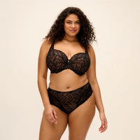 A woman modeling a black lace lingerie set consisting of a bra and matching high-waisted briefs against a plain beige studio background.
