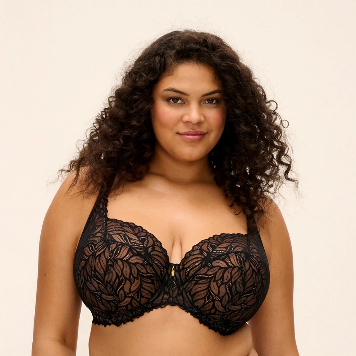 A woman with curly dark hair wearing a black lace underwire bra against a plain light beige studio background.