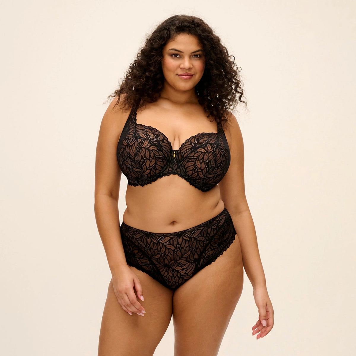 A woman modeling a black lace lingerie set consisting of a bra and matching high-waisted briefs against a plain beige studio background.
