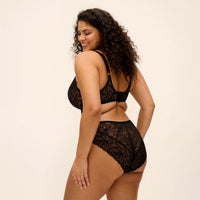 A woman modeling a black lace lingerie set, shown from a side-back angle against a plain off-white studio background.