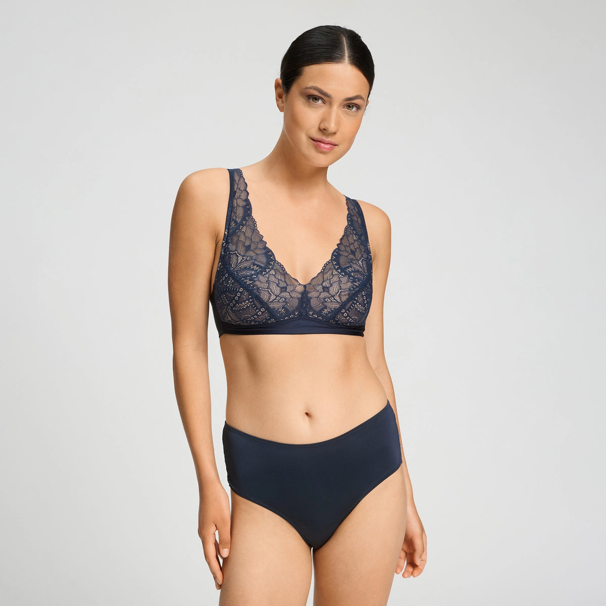 A woman modeling a two-piece lingerie set consisting of a navy blue lace bralette and matching solid navy high-waisted briefs, shot against a light gray studio background.