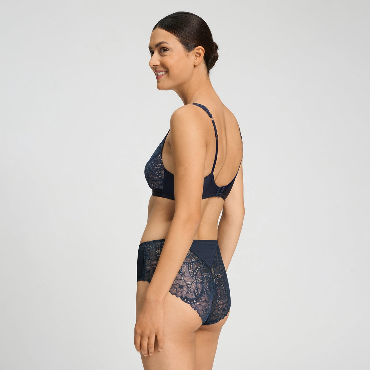 A woman modeling a navy blue lace bra and matching high-waisted briefs against a light gray studio background.