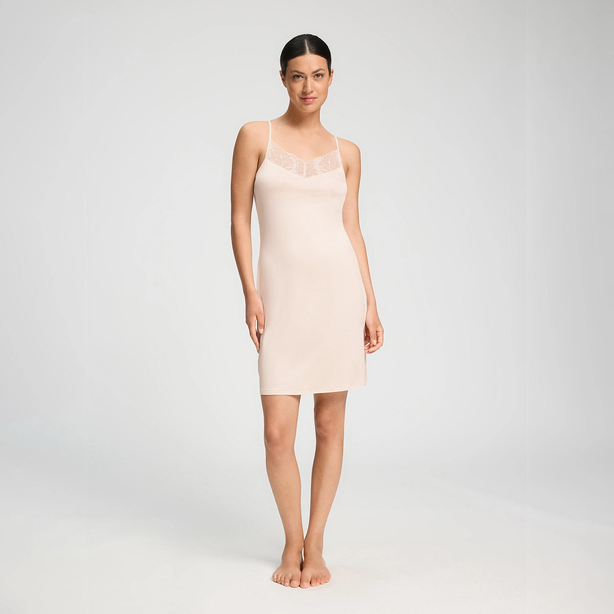 A woman modelling a light beige nightdress dress with thin straps and a lace V-neckline, standing against a plain light grey studio background.