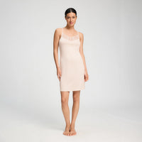 A woman modelling a light beige nightdress dress with thin straps and a lace V-neckline, standing against a plain light grey studio background.