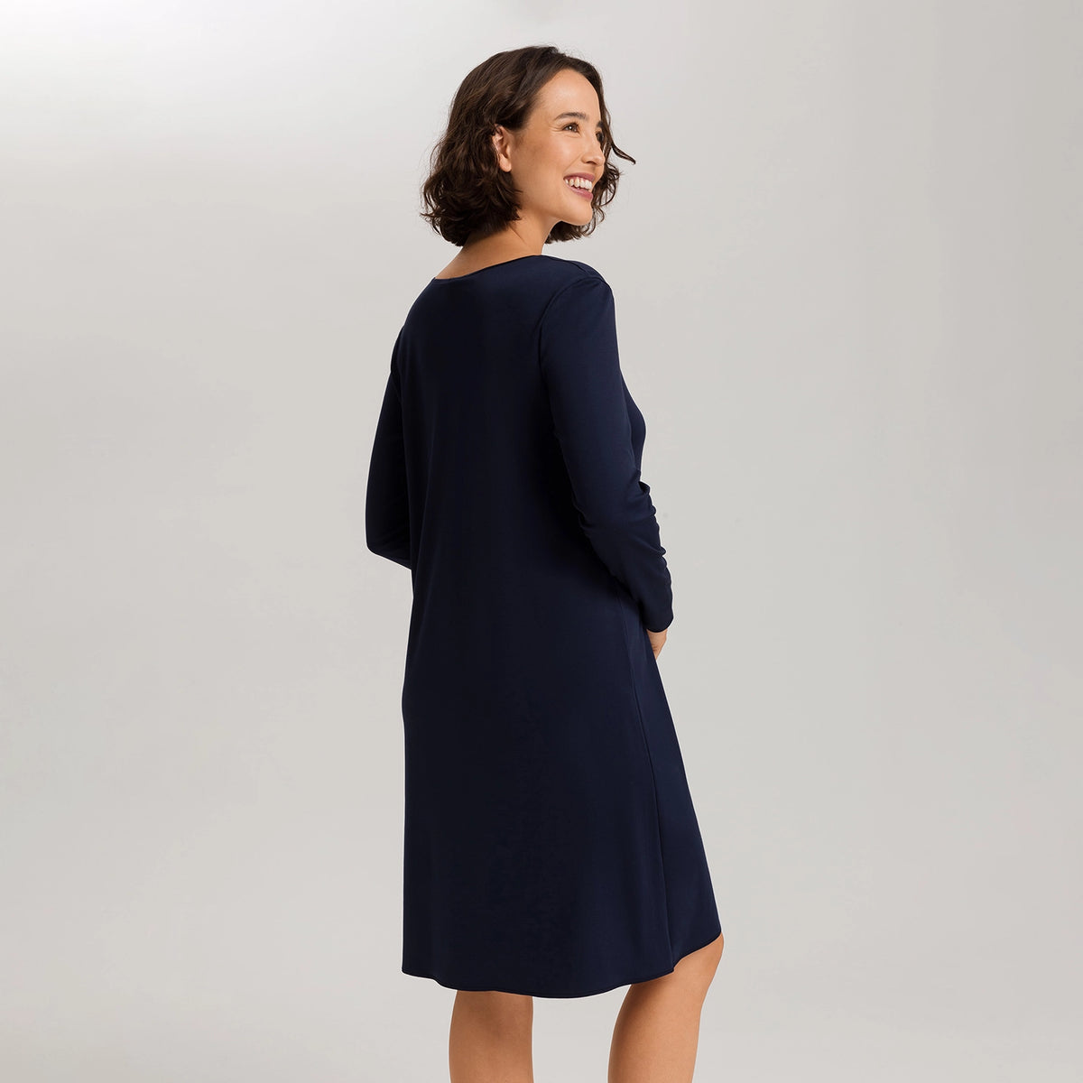 A woman wearing a dark navy blue long-sleeve shift dress, photographed from a three-quarters back angle against a light gray studio background.