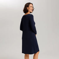 A woman wearing a dark navy blue long-sleeve shift dress, photographed from a three-quarters back angle against a light gray studio background.