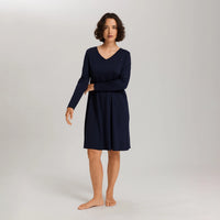 A woman modeling a long-sleeve V-neck knit dress in dark navy blue, standing barefoot against a light gray studio background.