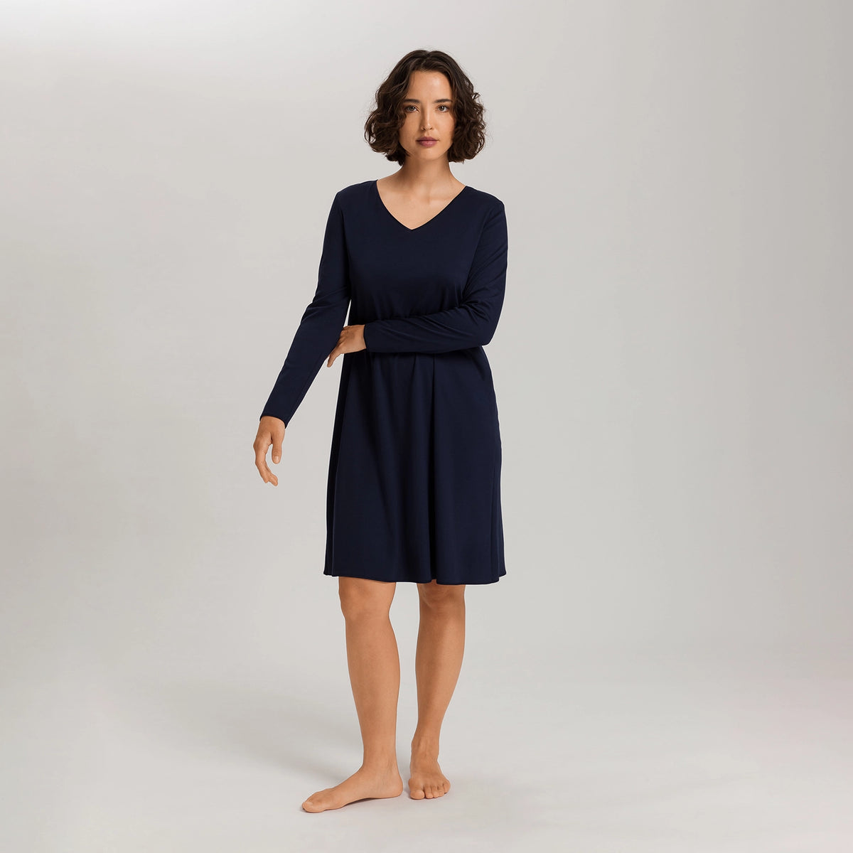A woman modeling a long-sleeve V-neck knit dress in dark navy blue, standing barefoot against a light gray studio background.