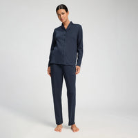 A woman modeling a matching navy blue pajama set consisting of a long-sleeve button-front top and straight-leg pants, photographed against a clean light gray studio background.