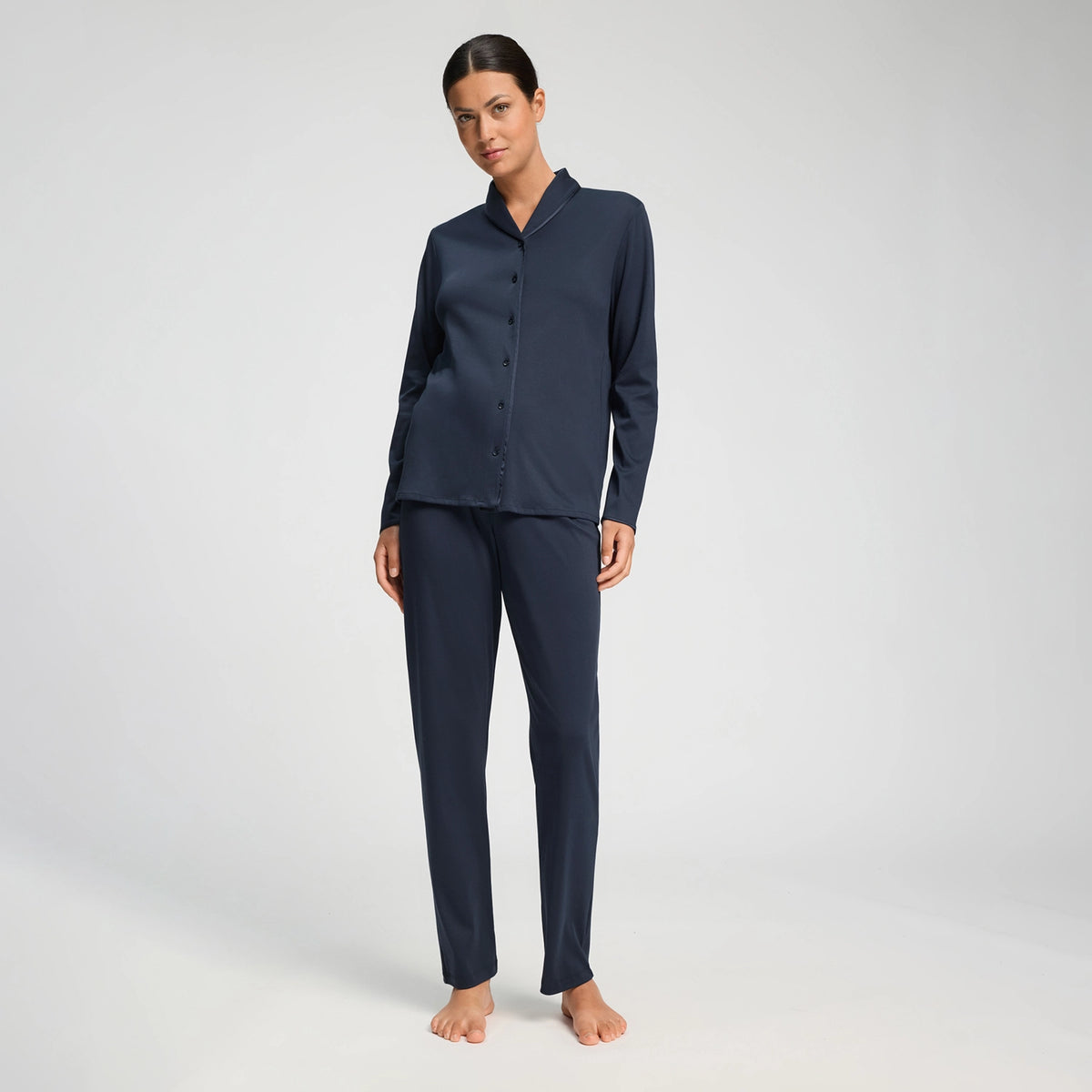 A woman modeling a matching navy blue pajama set consisting of a long-sleeve button-front top and straight-leg pants, photographed against a clean light gray studio background.