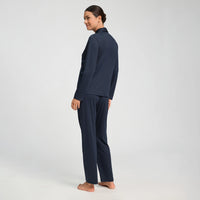 A woman stands barefoot in profile wearing a matching navy blue two-piece lounge set consisting of a long-sleeve top and wide-leg pants, photographed against a seamless light gray studio background.