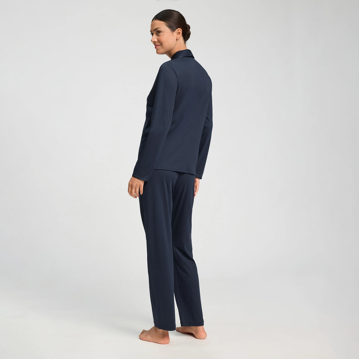 A woman stands barefoot in profile wearing a matching navy blue two-piece lounge set consisting of a long-sleeve top and wide-leg pants, photographed against a seamless light gray studio background.