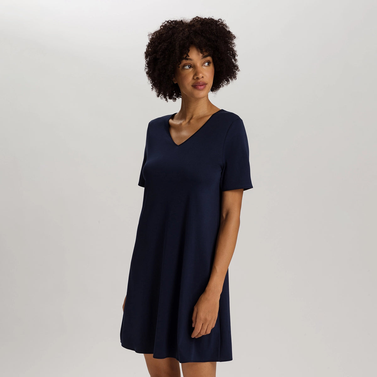 A woman with curly dark hair wearing a short-sleeve navy blue V-neck shift dress, standing against a light gray studio background.