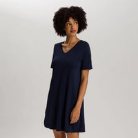 A woman with curly dark hair wearing a short-sleeve navy blue V-neck shift dress, standing against a light gray studio background.