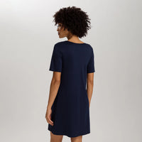 A woman with curly dark hair is shown from the back wearing a short-sleeve navy blue shift dress against a light gray studio background.