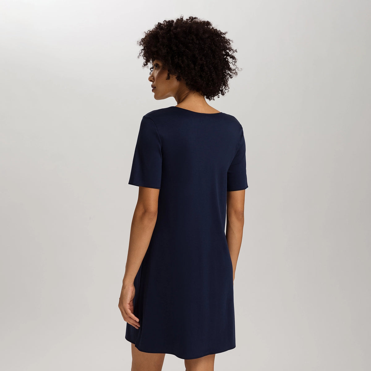 A woman with curly dark hair is shown from the back wearing a short-sleeve navy blue shift dress against a light gray studio background.