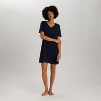 A woman with curly dark hair stands barefoot in a studio wearing a short-sleeve V-neck nightgown or tunic dress in dark navy blue against a light gray background.