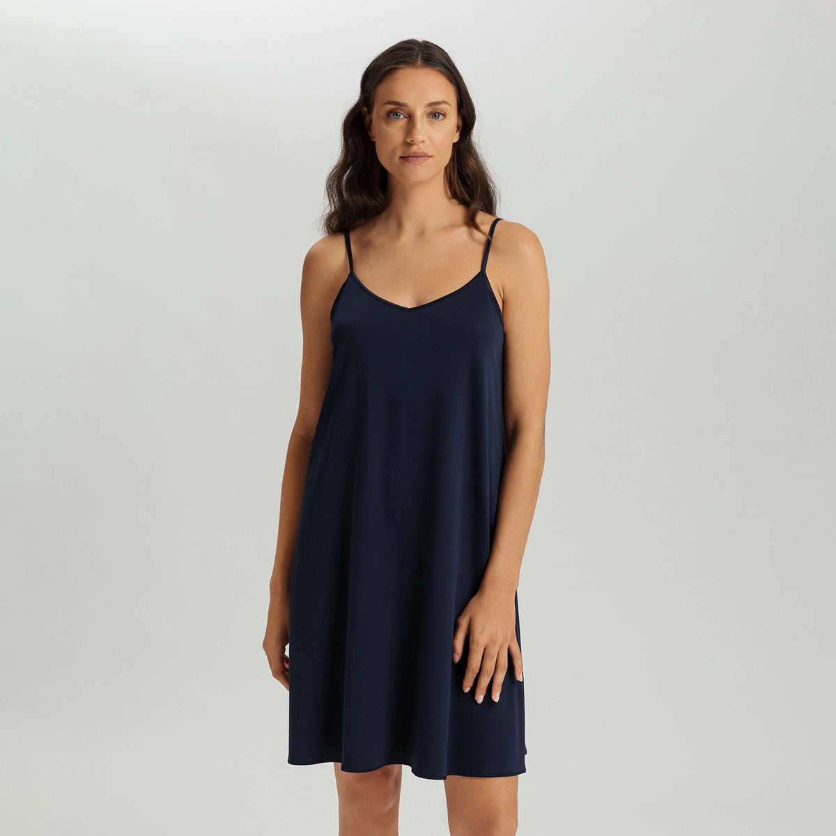A woman modeling a navy blue slip-style camisole dress with thin spaghetti straps and a V-neckline, photographed against a neutral light gray studio background.