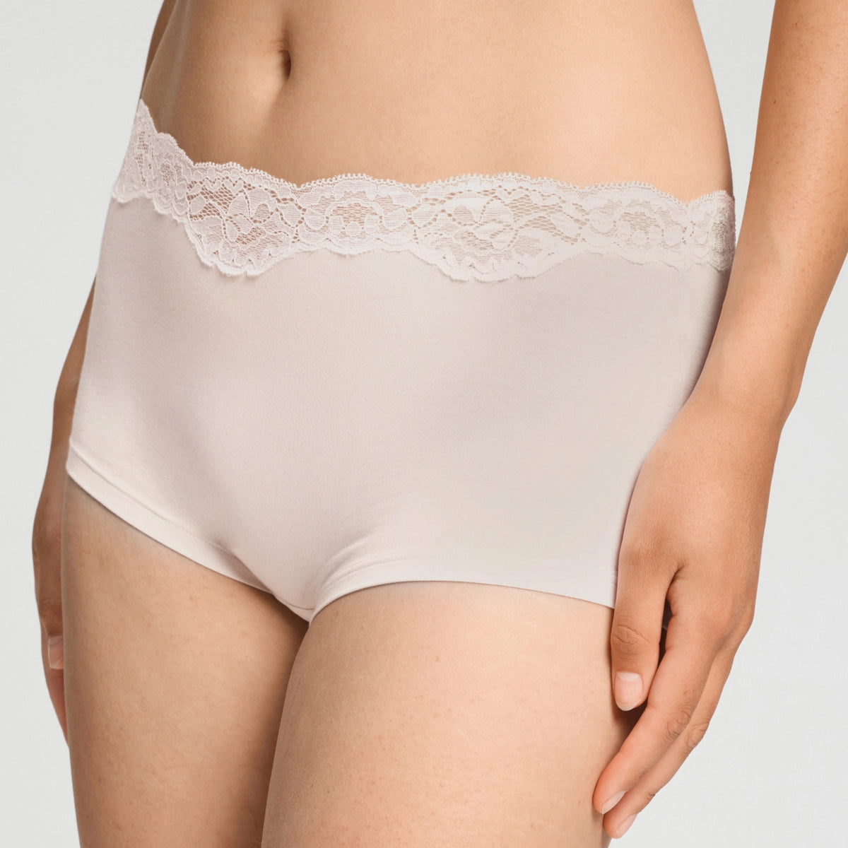 A close-up of a model wearing high-waisted, seamless briefs in a light beige color with a wide white lace waistband, set against a plain light gray background.