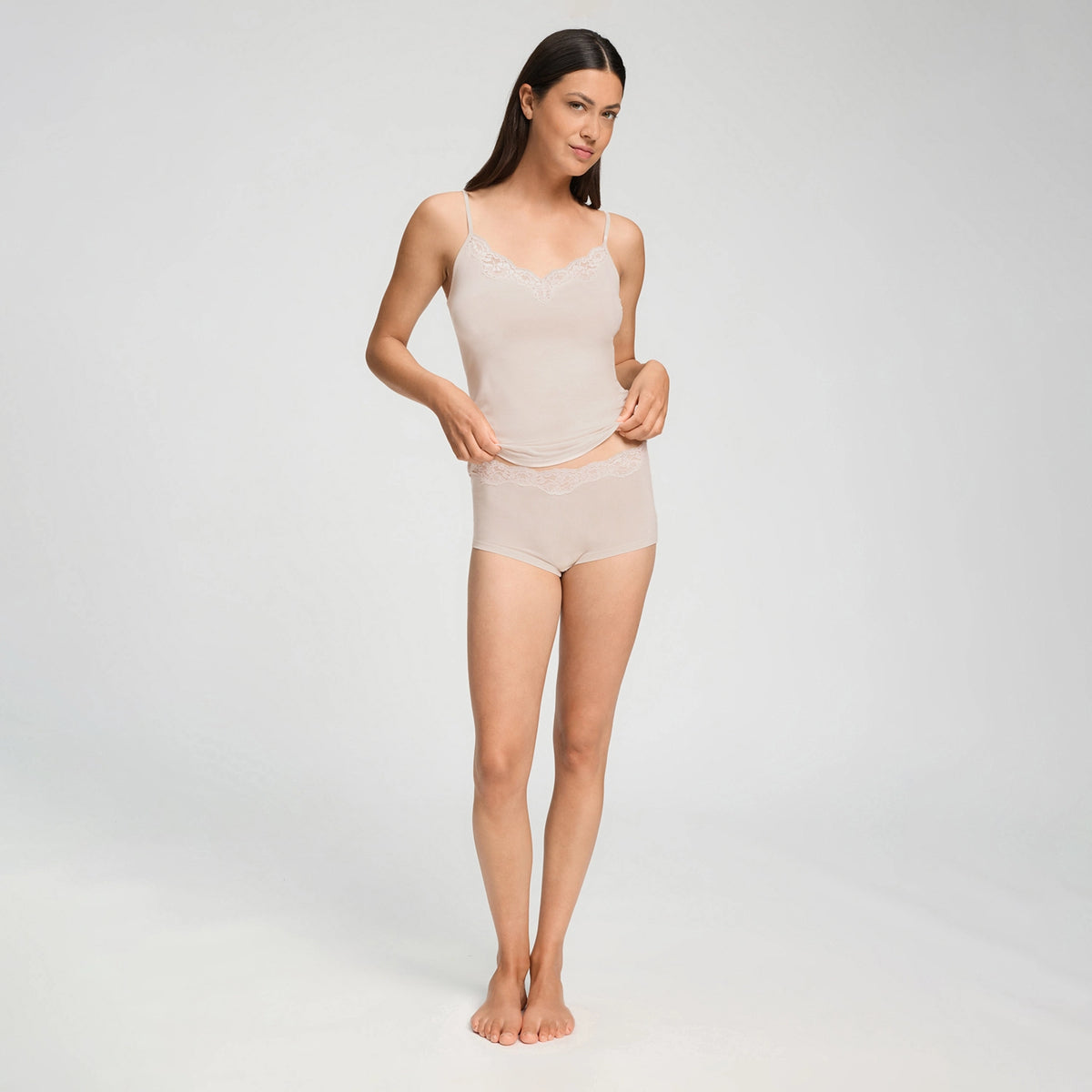 A woman modeling a light beige two-piece lingerie set consisting of a camisole top with lace trim and matching briefs, photographed against a seamless light gray studio background.