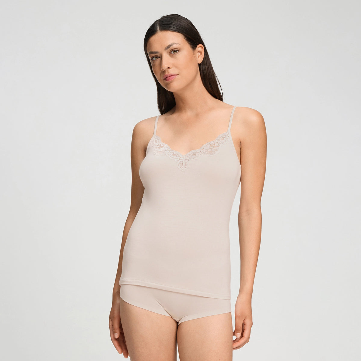 A woman modeling a light beige camisole top with delicate lace trim at the neckline, paired with matching high-waisted briefs, against a plain light gray studio background.