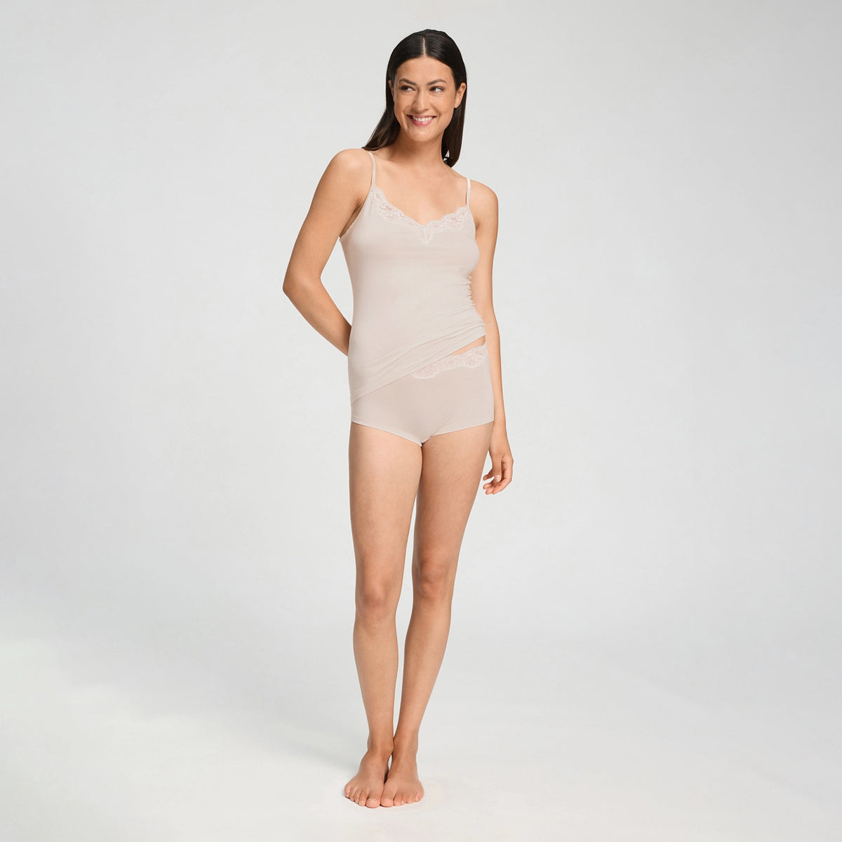 A woman modeling a light beige sheer lingerie bodysuit with lace trim, standing against a plain light gray studio background.