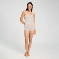 A woman modeling a light beige sheer lingerie bodysuit with lace trim, standing against a plain light gray studio background.