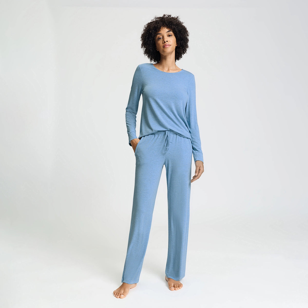 A woman modeling a light blue two-piece loungewear set consisting of a long-sleeve crewneck top and matching drawstring pants, standing against a plain white studio background.