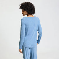 A woman with curly dark hair is shown from the back wearing a light heather blue long-sleeve top and matching pants against a plain light gray studio background.