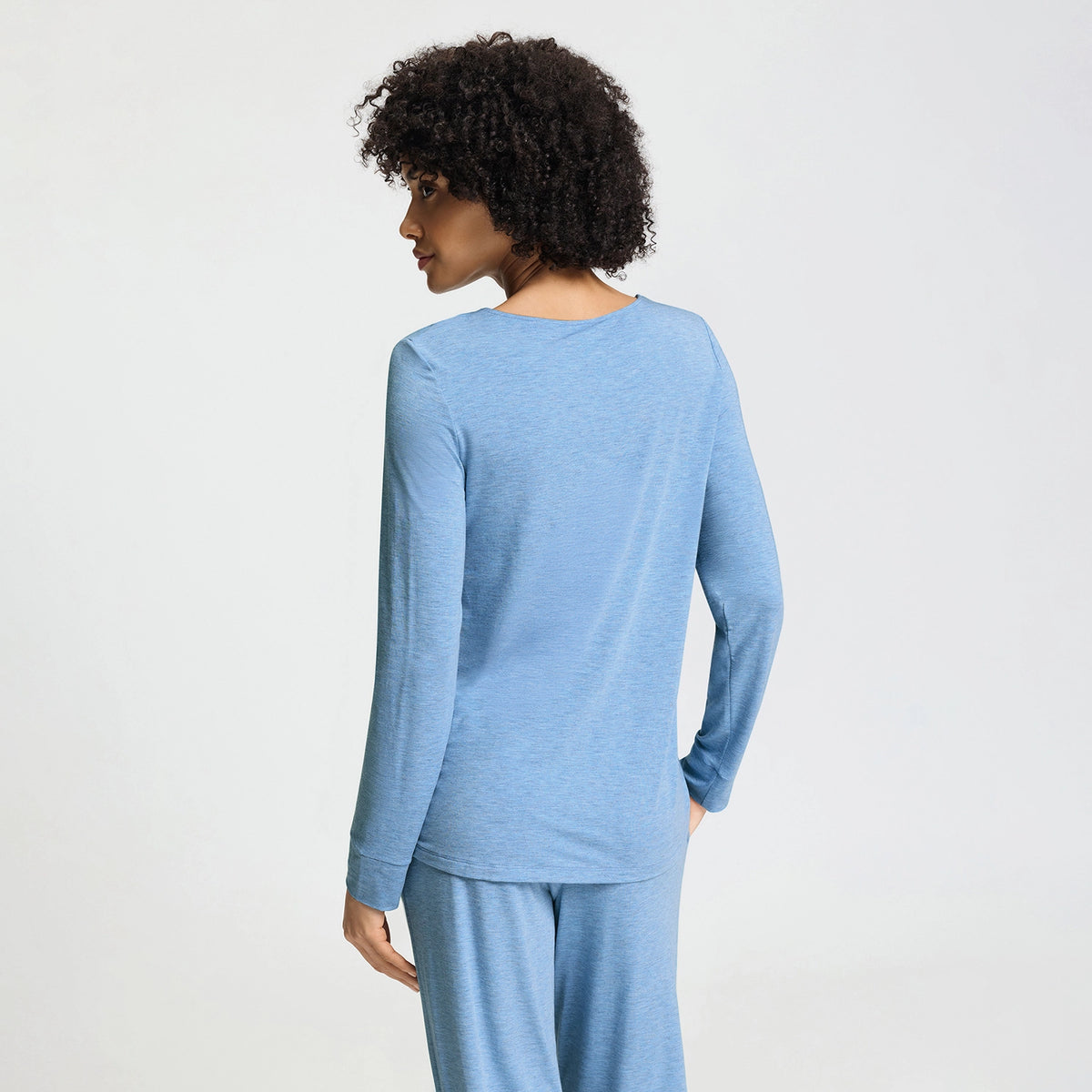 A woman with curly dark hair is shown from the back wearing a light heather blue long-sleeve top and matching pants against a plain light gray studio background.