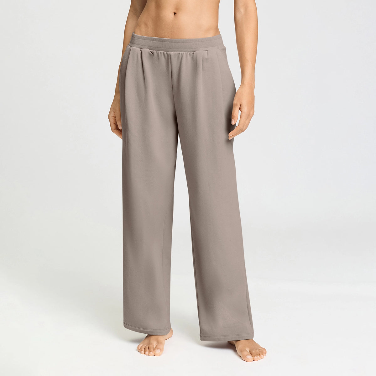 A model wearing wide-leg lounge pants in a taupe-gray color, standing barefoot against a clean white studio background.