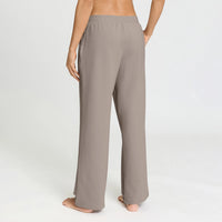A person shown from the back wearing loose, wide-leg lounge pants in a warm taupe-gray color, standing barefoot against a clean white studio background.