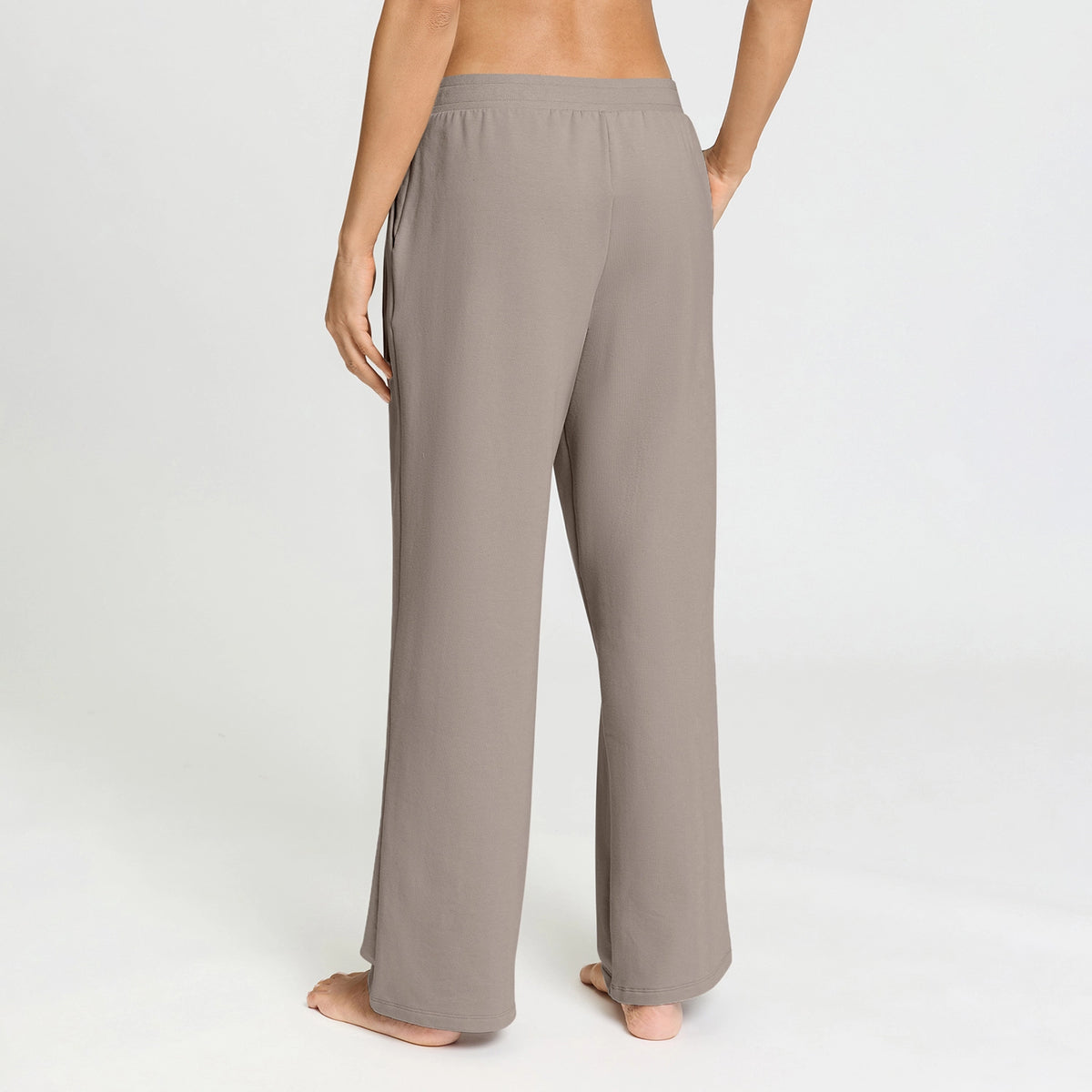 A person shown from the back wearing loose, wide-leg lounge pants in a warm taupe-gray color, standing barefoot against a clean white studio background.