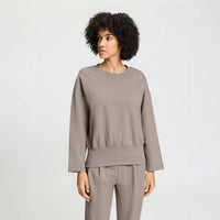 A woman with curly black hair wearing a taupe long-sleeve cropped sweatshirt and matching pants, photographed against a light gray studio background.