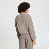 A model with curly dark hair is shown from the back wearing a taupe-colored long-sleeve top and matching pants against a light gray studio background.