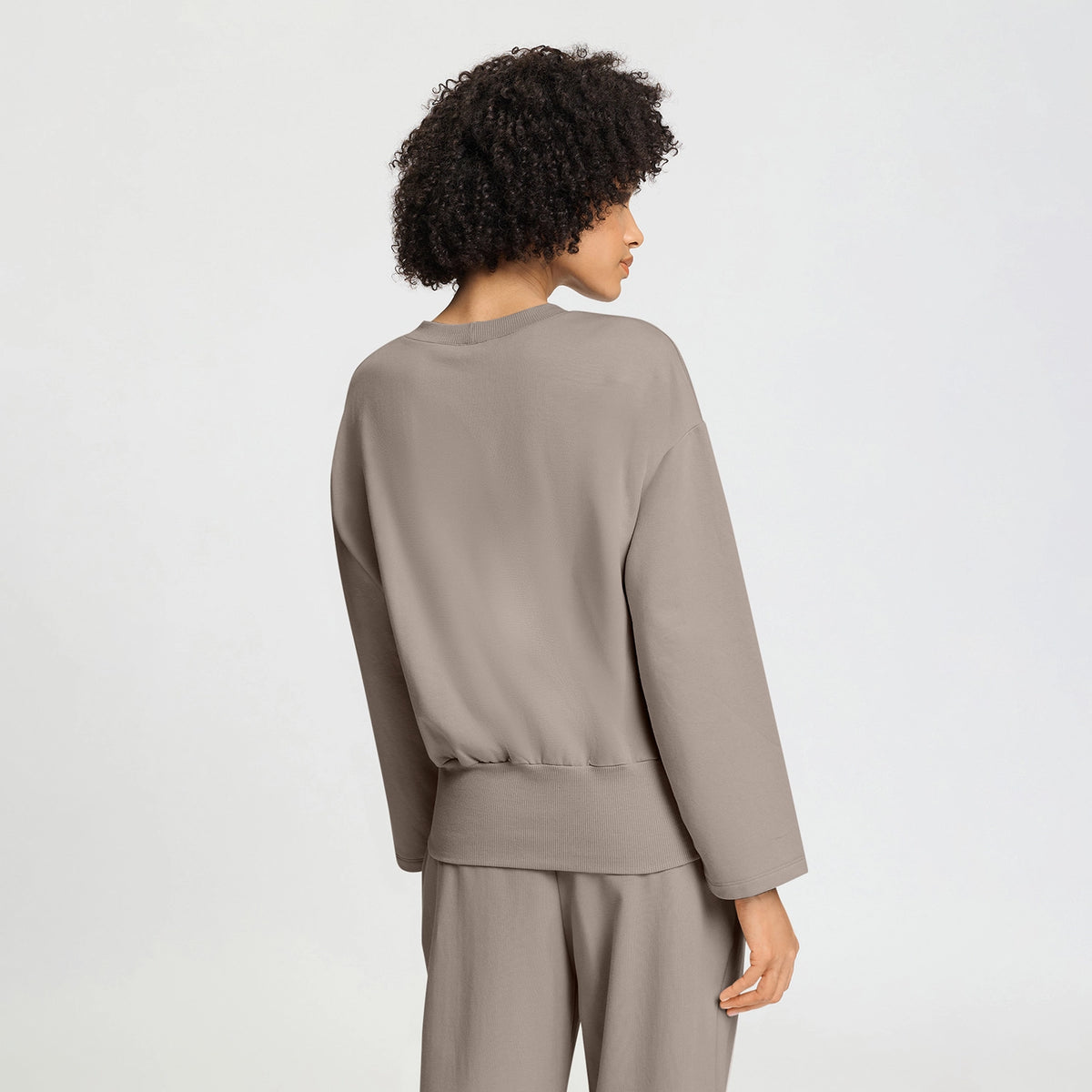 A model with curly dark hair is shown from the back wearing a taupe-colored long-sleeve top and matching pants against a light gray studio background.