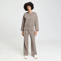 A woman modeling a matching taupe two-piece lounge set consisting of a long-sleeve top and wide-leg trousers, paired with white slip-on shoes, against a light gray studio background.