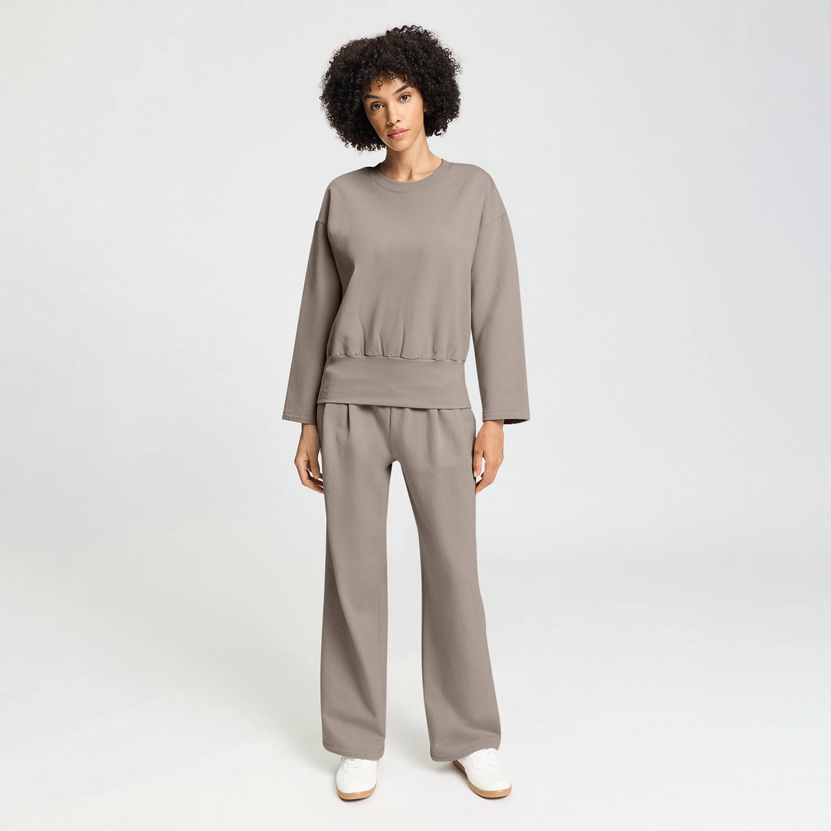 A woman modeling a matching taupe two-piece lounge set consisting of a long-sleeve top and wide-leg trousers, paired with white slip-on shoes, against a light gray studio background.