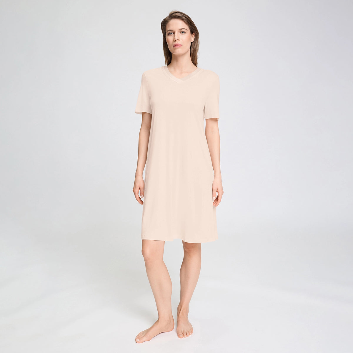 A woman modelling a short-sleeve V-neck nightgown in a light beige colour, standing against a plain light grey studio background.