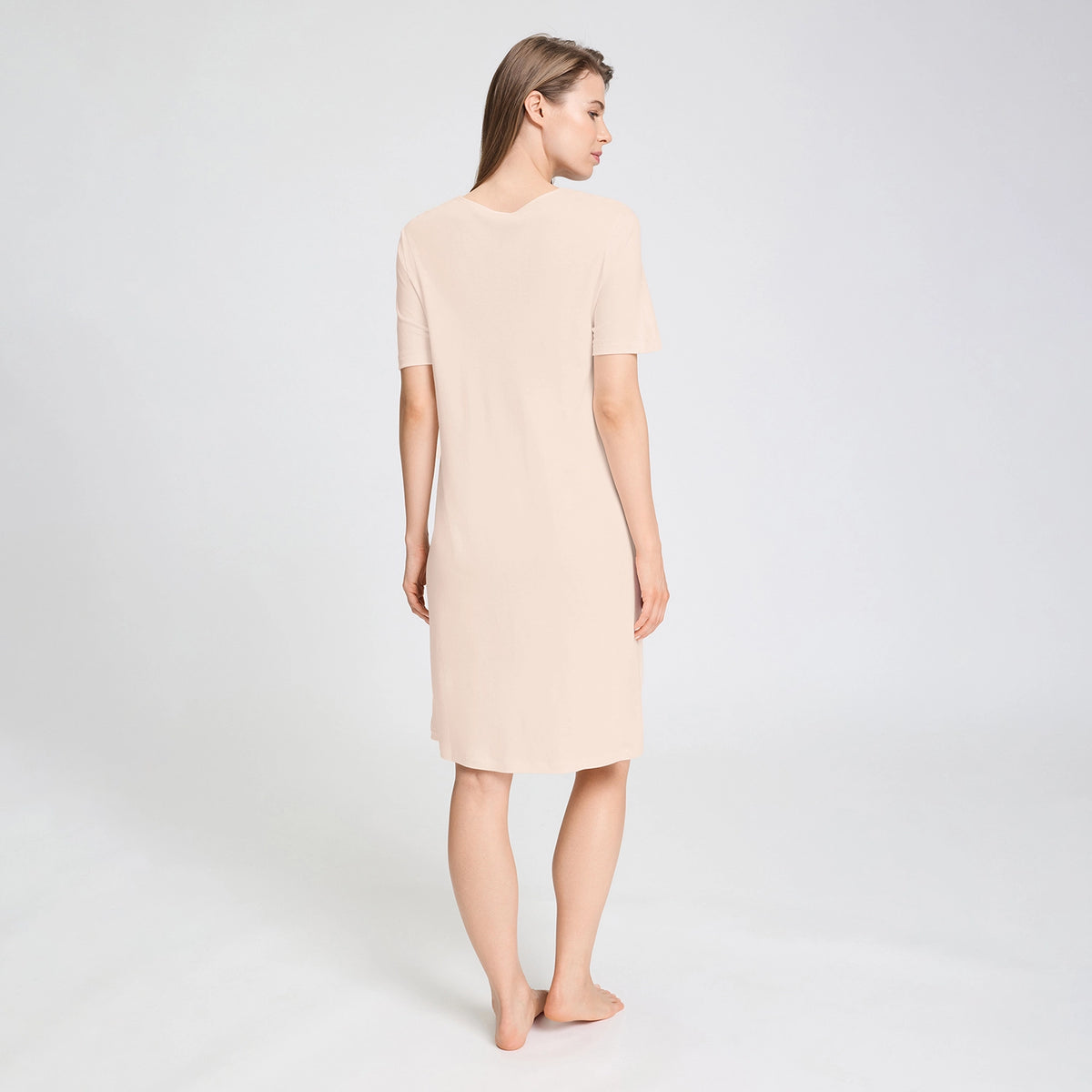 A back view of a woman modelling a short-sleeve V-neck nightgown in a light beige colour, standing against a plain light grey studio background.