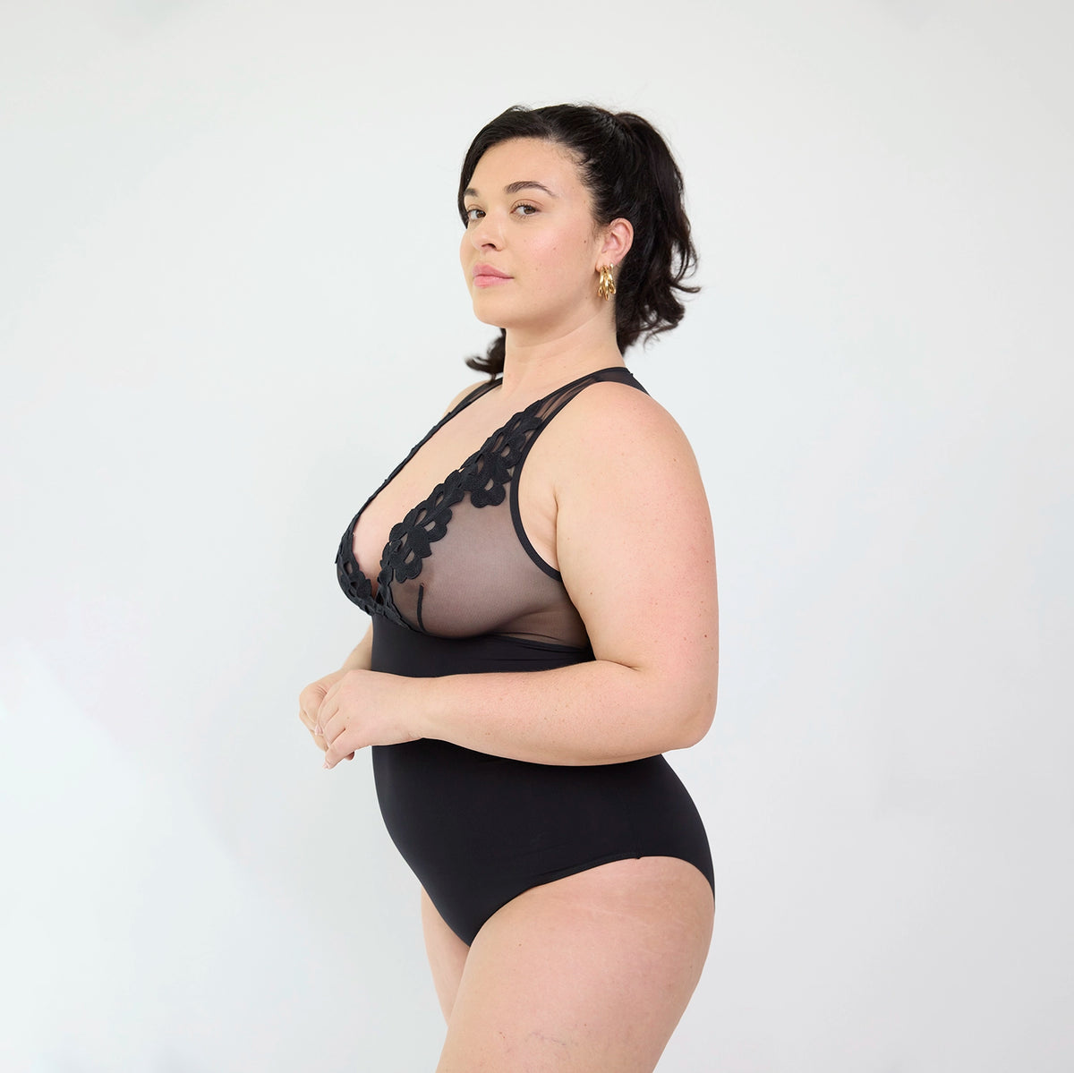 A woman modeling a black sheer mesh and lace bodysuit against a plain light gray studio background.