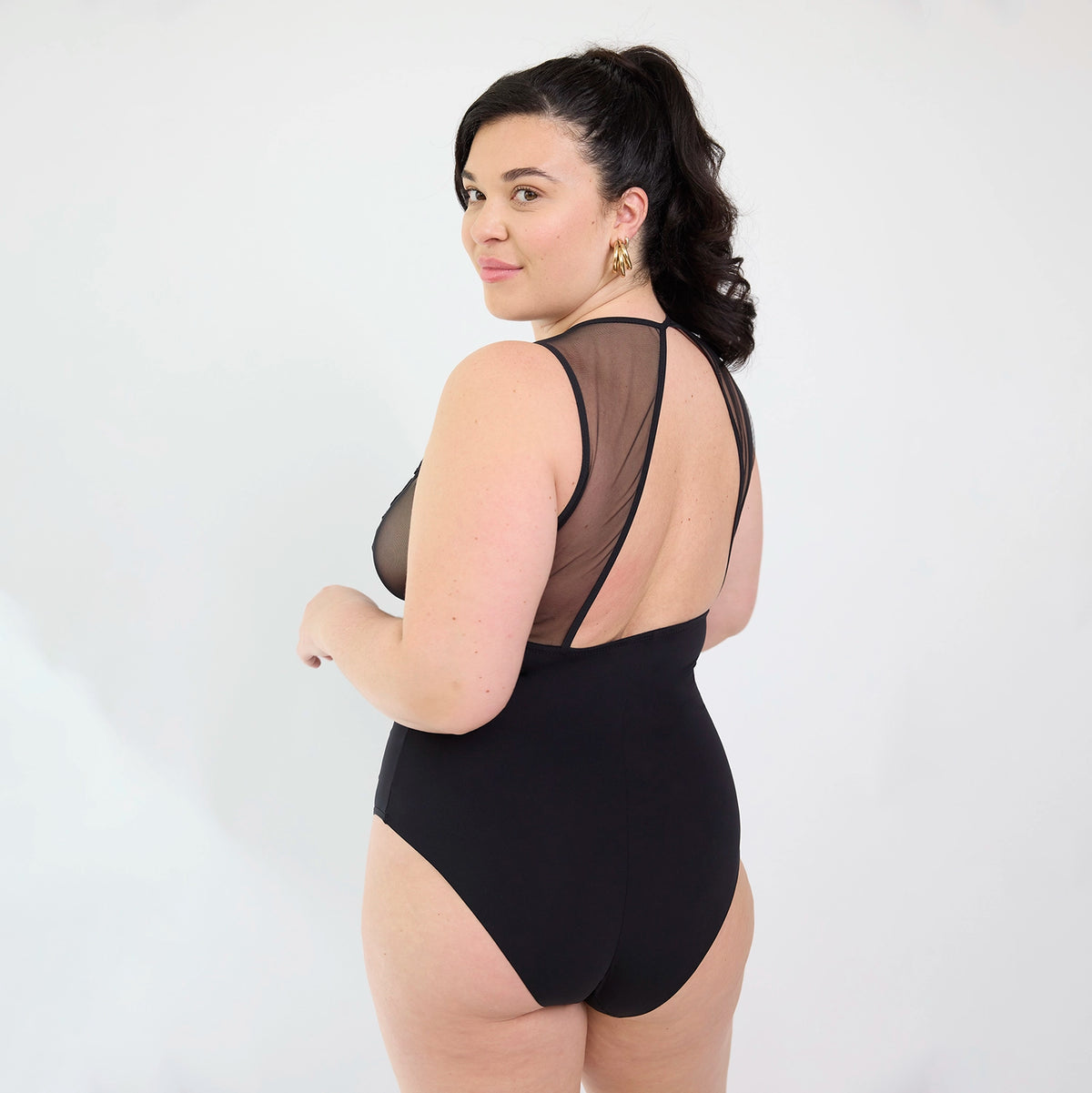 A woman modeling a black one-piece bodysuit with sheer mesh shoulder panels and an open back, photographed against a plain light background.