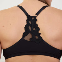 Close-up back view of a black sports bra with an intricate floral lace and mesh back strap design, shown on a model against a light background.