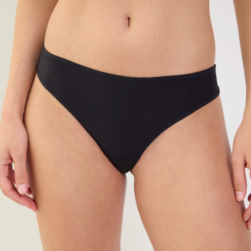 A close-up of a model wearing a solid black bikini bottom against a clean, light off-white background.
