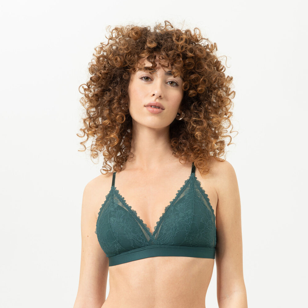 A woman with curly brown hair is shown from the front wearing a dark green bralette with thin straps and lace trim, against a plain white studio background.